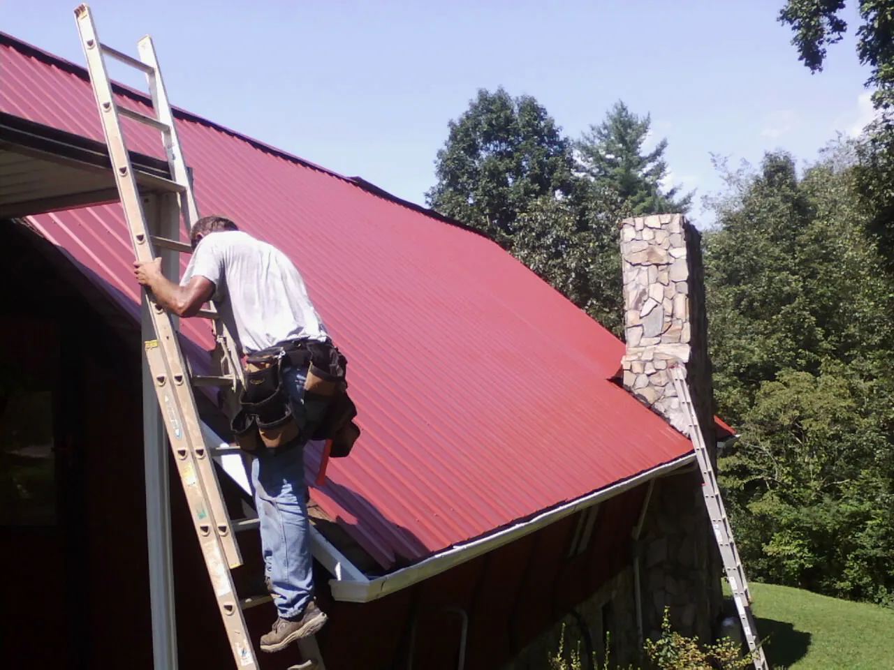 Roofer on a ladder beside a red metal roof and stone chimney — Soffit & Fascia Repair by Roofing Brothers in Westerville